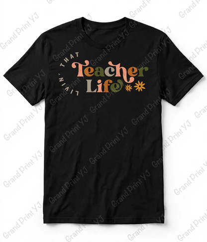 A black short-sleeve crewneck t-shirt with a colorful graphic on the chest that reads 'THAT Teacher Life**'. The word 'Teacher' is written in a wavy orange font, 'Life' in a green font, and the remaining text is in a light beige font.