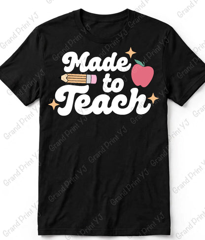 A black short-sleeve crewneck t-shirt with a 'Made to Teach' graphic on the chest. The graphic features white cursive text, a pink apple with a green stem, a yellow pencil, and small gold stars.
