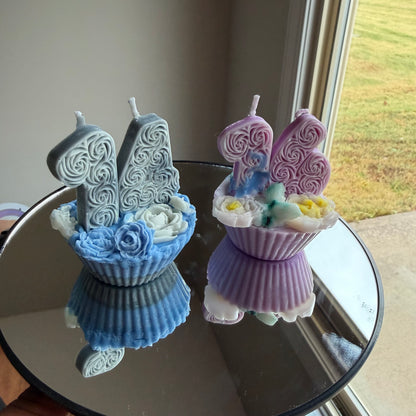 Two decorative candles shaped like cupcakes with candles on a reflective surface.