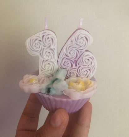 Hand holding a decorative cupcake-shaped candle with floral details against a plain background